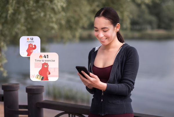 En İyi Çevrimiçi Kişisel Koç. Göl kenarındaki bir korkulukta duran genç kadın, kırmızı ayı maskotu ve alevler gösteren iki yüzen fitness uygulaması bildirim kartı ile akıllı telefonuna bakıyor