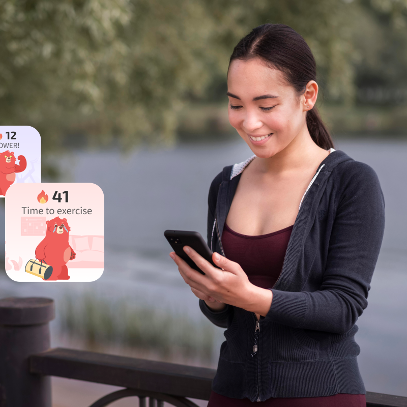 Najlepszy trener osobisty online. Młoda kobieta stojąca przy balustradzie nad jeziorem, patrząca na swój smartfon z dwiema pływającymi kartami powiadomień aplikacji fitness przedstawiającymi maskotkę czerwonego niedźwiedzia, płomienie
